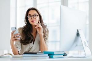 Female company office worker in formal wear uses modern gadget for work, poses at workplace near monitor, plans working schedule, browses information online, poses against window. Working time photo