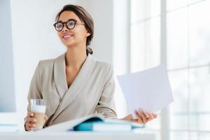 Cheerful businesswoman reads paper documents, focused aside with smile drinks milkshake sits at desktop makes business plan for project. Female executive manager in spectacles poses in coworking space photo