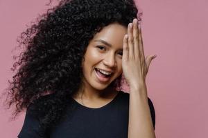 Happy teenage girl has fun and hides face with palm, has joyful expression, curly bushy hair, dressed in casual black clothes, isolated on pink background. Shy glad young woman gets compliment photo