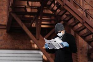 el hombre concentrado usa gafas de sol y abrigo, máscara médica, guantes de goma protectores, lee noticias recientes sobre la propagación del coronavirus del periódico, posa contra un fondo borroso con escaleras. foto