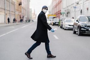 Virus covid -19. Man crosses road, dressed in black coat, hat and sunglasses, wears ptotective mask for pandemic virus protection, poses outdoor at busy street with many transport and people photo