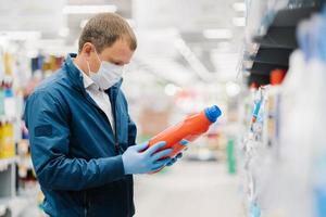 Man makes shopping in supermarket, holds bottle of washing detergent, reads product information, wears protective medical mask and gloves during coronavirus outbreak, chooses something for household photo