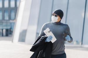 Shot of man walks outdoor, protects from flu, coronavirus spreading, drinks coffee, looks aside with pensive expression, reads newspaper, follows quarantine rules. 2019-ncov. Pandemic virus. photo