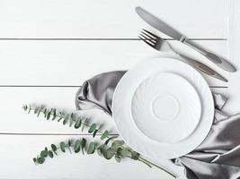 top view of simple white table setting with silk napkin and green leaves. wooden table. mock up for dinner meal. restaurant menu. photo