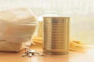 a medical mask over a packet of corn grits ,a tin of canned food ,some pasta and the keys to the house on a wooden background by the window photo