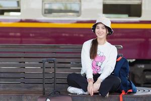 Asian beautiful woman in white long sleeved in a hat sits smiling on chair with her bag at train platform in social distancing as a new normal lifestyle.