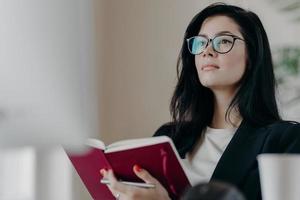 Beautiful woman with thoughtful expression, holds opened notepad, writes time management, makes plans for productive day, wears eyeglasses, poses indoor. Self employed female writes down ideas photo