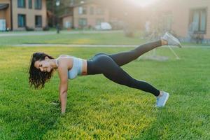 una mujer deportiva feliz se para en una pose de tablón, levanta las piernas y hace ejercicios físicos para mantenerse en forma, se viste con la parte superior y las polainas, practica deporte regularmente, disfruta de un buen día de verano, posa en la hierba verde foto