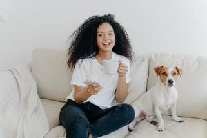 Delighted dark skinned woman poses in modern apartment, sits on comfortable sofa with pet, drinks coffee, uses mobile phone for online communication, being in good mood, flipping news, uses app photo
