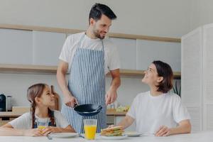 Happy father, mother and daughter gather together at kitchen, eat delicious breakfast, dad prepared fried eggs, being in good mood, ready to start new day. Lovely family enjoy tasty meal at home photo