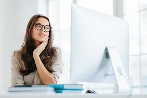 Indoor shot of serious brunette lady focused in monitor of computer, uses wireless internet for publication, keeps hand on cheek, has attentive look, sits at desktop, works on business contract photo