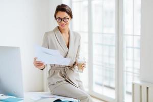 Pleased dark haired woman with smile, checks bills in documents, dressed beige business suit, drinks beverage, sits at desktop near big monitor, holds paper sheet with charts, poses near window photo