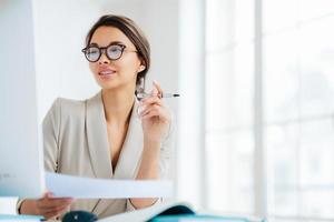 Prosperous businesswoman focused in monitor of computer, studies financial issues, holds document and pen, works in office, wears formal clothing and spectacles, poses over white background. photo