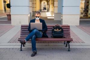 Photo of busy man freelancer in mask does distance work, keyborads on laptop computer, sits at wooden bench outside, avoids crowded places during quarantine time. Traveling and coronavirus concept
