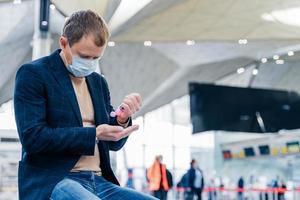 Photo of man in medical face mask, uses disinnfectant spray for hands, protects from coronavirus in crowded place, poses in airport, waits for flight. Health care concept. Sanitation, virus prevention