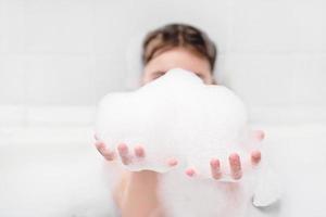 girl playing with bath foam in the bathroom. little girl making froth and holding it in hands. kids hygiene, evening routine photo