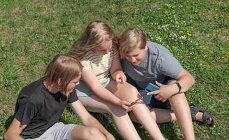 group of teenagers looking at smartphone screen and smiling and chatting. teens sitting on a lawn grass wearing casual clothes spending time outdoors photo