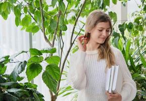 teenager girl holding school books and smiling. back to school concept. teen girl wearing casual white pullover looking aside. large green plants aside photo