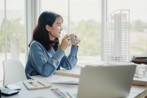 enfoque selectivo, joven arquitecto o ingeniero asiático bebiendo té, café durante un descanso en la planificación de los planos de las estructuras de construcción. proyectos de diseño de arquitecto o ingeniero en la oficina. foto