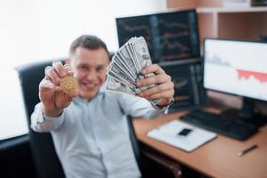 concepto de éxito. hombre de negocios con bitcoin y dinero en las manos mientras está sentado en una oficina moderna con muchos monitores con gráficos foto