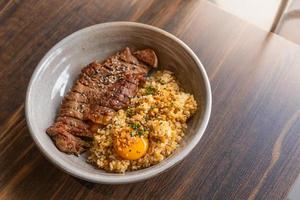 arroz frito con ajo y grasa de ternera con bistec de ternera y cubierta con yema de huevo en escabeche en un bol sobre una mesa de madera. carnes a la brasa es menú es comida fusión foto