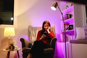 Relaxed girl using phone in the night sitting on an armchair in the living room at home photo
