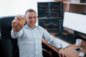 Shows physical sample. Businessman holding bitcoin in hand while sitting in modern office with many monitors with graphs photo