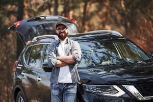 Bearded man near his brand new black car in the forest. Vacations concept photo