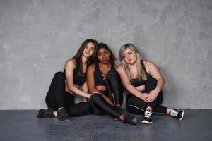 Brunette leaning on the black girl. Group of multi ethnic women sitting in the studio against grey background photo