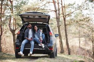 There is only two of us. Sitting on rear part of automobile. Enjoying the nature. Couple have arrived to the forest on their brand new black car photo