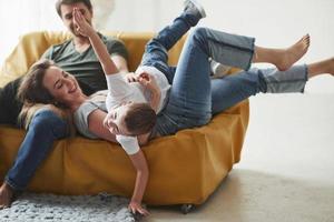 Kid imagine that he is airplane. Happy family have fun on the yellow sofa in the living room of their new house photo