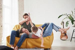 Kid lying on his parents. Happy family have fun on the yellow sofa in the living room of their new house photo