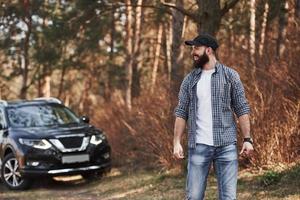 Man and nature. Bearded man near his brand new black car in the forest. Vacations concept photo