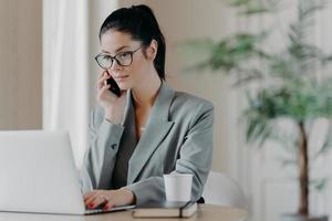 Serious brunette woman keyboards information, concentrated into laptop computer, dressed in formal outfit, poses at coworking space, works remotely, involved in working process during daytime. photo
