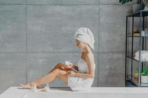 Horizontal shot of relaxed young European woman applies moisturizing body cream on legs, wrapped in bath towel, has tender smile, healthy refreshed skin after taking bath, poses in cozy bathroom. photo