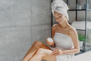 foto de una joven europea sensual que usa crema corporal después de bañarse cuida su piel envuelta en toallas blancas posa contra el fondo del baño. concepto de bienestar, mimos y bienestar