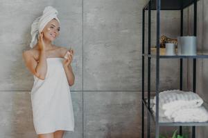 una joven europea complacida se aplica loción facial sostiene una botella de producto cosmético envuelta en una toalla de baño blanca y se apoya contra la pared gris en el baño. concepto de belleza y limpieza de cosmetología de personas foto