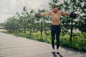 Photo of athletic strong fitness man works out with barbells, demonstrates power and energy, leads active lifestyle, shows strength, motivation, does biceps lifting, has physical workout outdoor