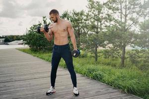 Sport and bodybuilding concept. Muscular bearded European man raises heavy barbells wears sport trousers and sneakers flexes muscles outdoors, does weightlifting exercises, leads active lifestyle photo