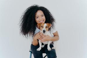 Smiling African American woman poses with domestic animal, looks gladfully at camera, cuddles dog, sits on chair, isolated over white background. Two family members. Homely relax. Forever together photo