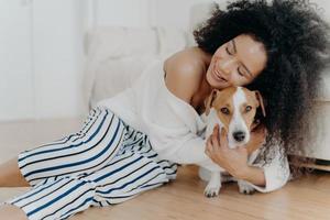 Affectionate young woman hugs dog with love and care, keeps eyes closed from pleasure, smiles gently, has healthy dark skin, poses on floor, petting animal. People, friendship and pets concept photo