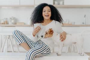 People, leisure and animals concept. Joyful dark skinned woman petts favourite dog, drinks hot beverage from white cup, looks happily aside spend time in kitchen. Caring host with jack russell terrier