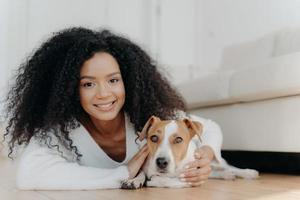 Pretty girl with Afro hair, lies on floor with dog, expresses pleasant emotions, poses in living room near couch, bought pet in new apartment. Woman host with beloved animal at home, share good moment photo
