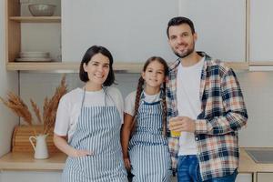 familia feliz pasar un buen rato juntos, posar en la cocina moderna en casa. un hombre alegre con camisa a cuadros sostiene un vaso de jugo, un niño pequeño con coletas, una linda ama de casa con delantal. niña con padres foto