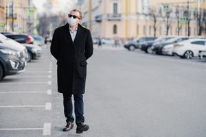 Full length shot of serious man walks at street, dressed in elegant outfit, wears medical mask to prevent coronavirus or another type of virus, stays safe during quarantine time. Pandemic situation photo