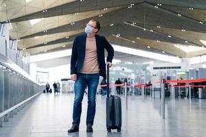 Full length shot of man passeger with suitcase in international airport wears protective face mask to avoid coronavirus infection. Male with luggage going to travel abroad. Epidemic, health care photo