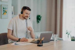 European male student learns foreign languages online has video lesson uses modern headset focused at laptop screen computer engaged in conference listens lecture attentively makes notes in diary photo