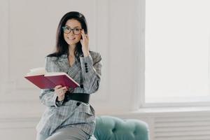 Smiling female business reporter in formal clothes holds opened notebook and pen, writes down information or checklist, wears formal clothes, works remotely from home, prepares for formal meeting photo