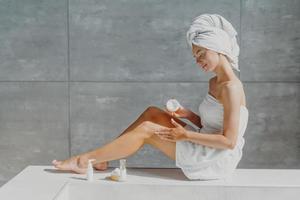 Horizontal shot of pretty woman with slender legs puts moisturising cream on healthy skin enjoys beauty routine after taking bath poses in bathroom wrapped in towel enjoys softness after lotion photo