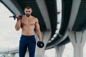 Outdoor shot of strong muscular man raises barbells and trains muscles stands under bridge has aim to have healthy body shape, has fitness training or physical workout. Athletic bodybuilder. photo